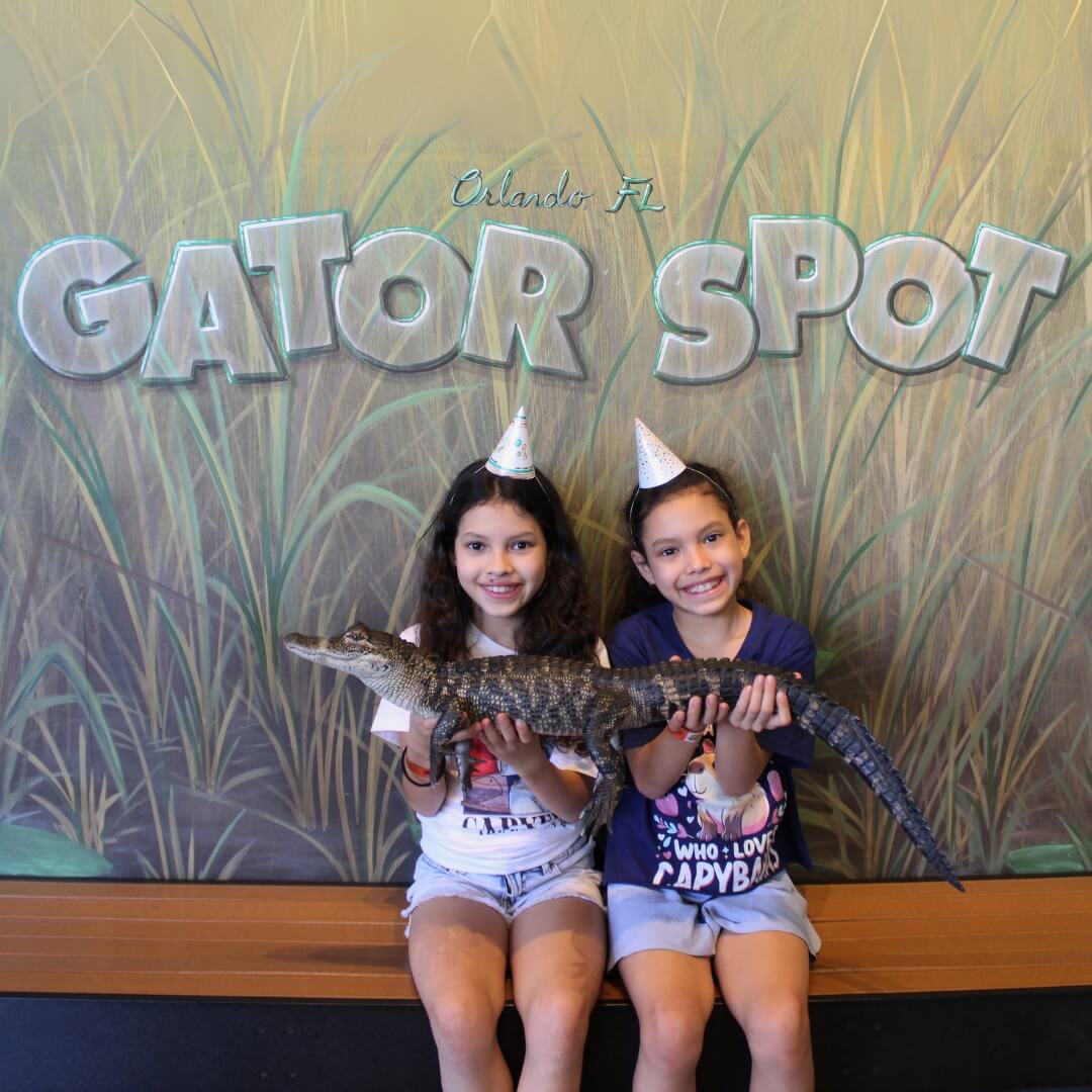 1 Two young girls wearing party hats hold a small alligator in front of a "GATOR SPOT" sign